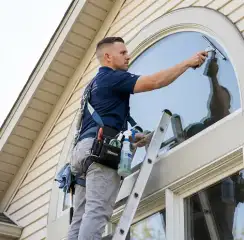 Professional window cleaner on a ladder using a squeegee to clean an exterior arched window, Little Rock, AR.